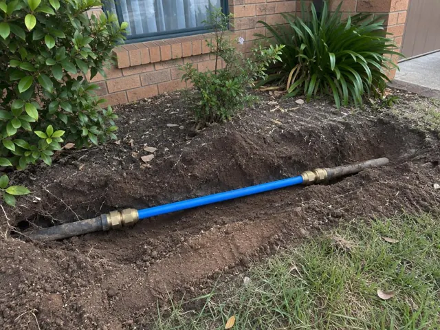 Blue repaired water pipe in an outdoor trench beside a brick house and garden in Penrith